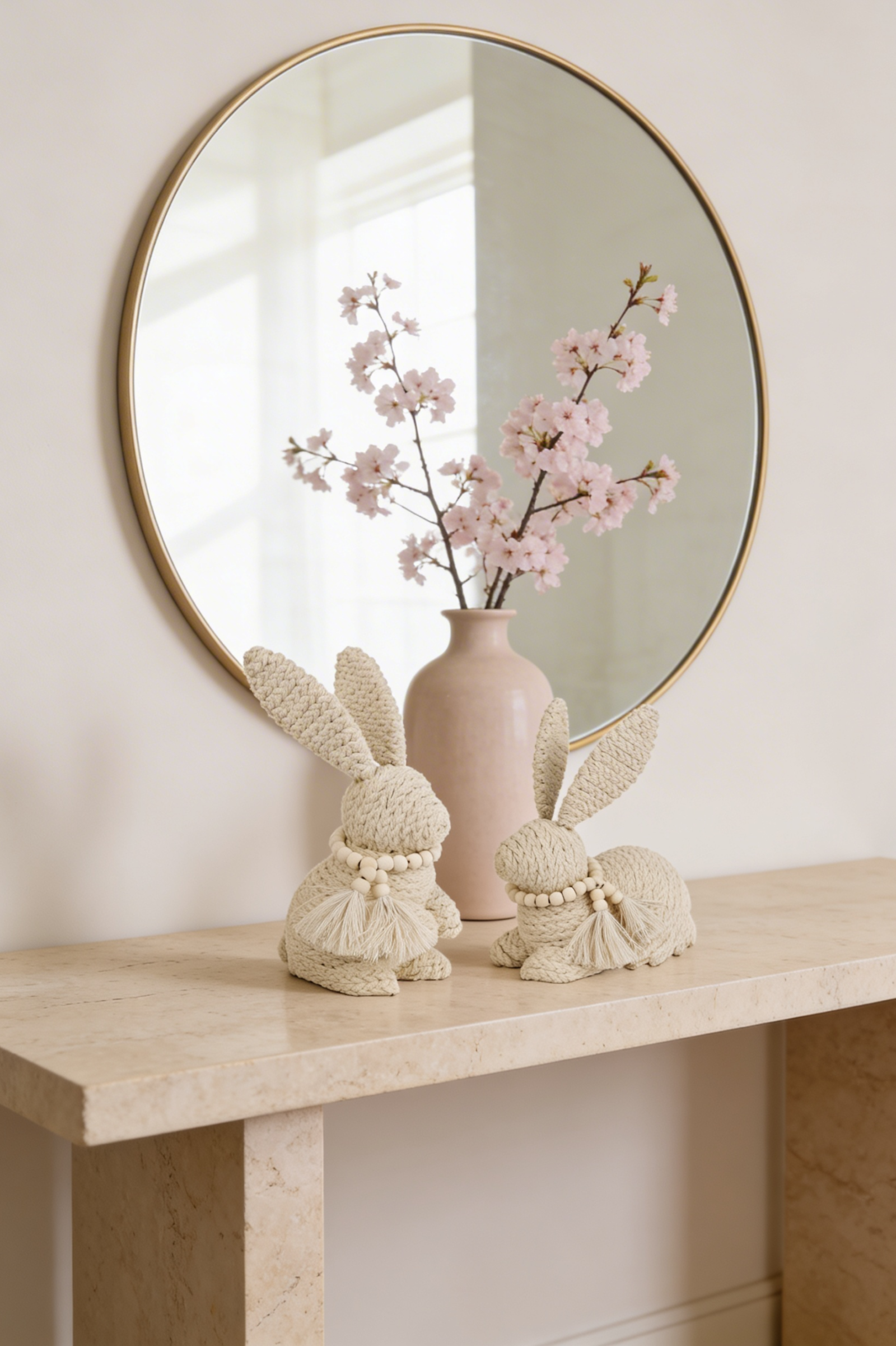 Two macramé rope bunny figurines with tassel and bead necklaces on a marble console beside a pink cherry blossom vase and gold mirror