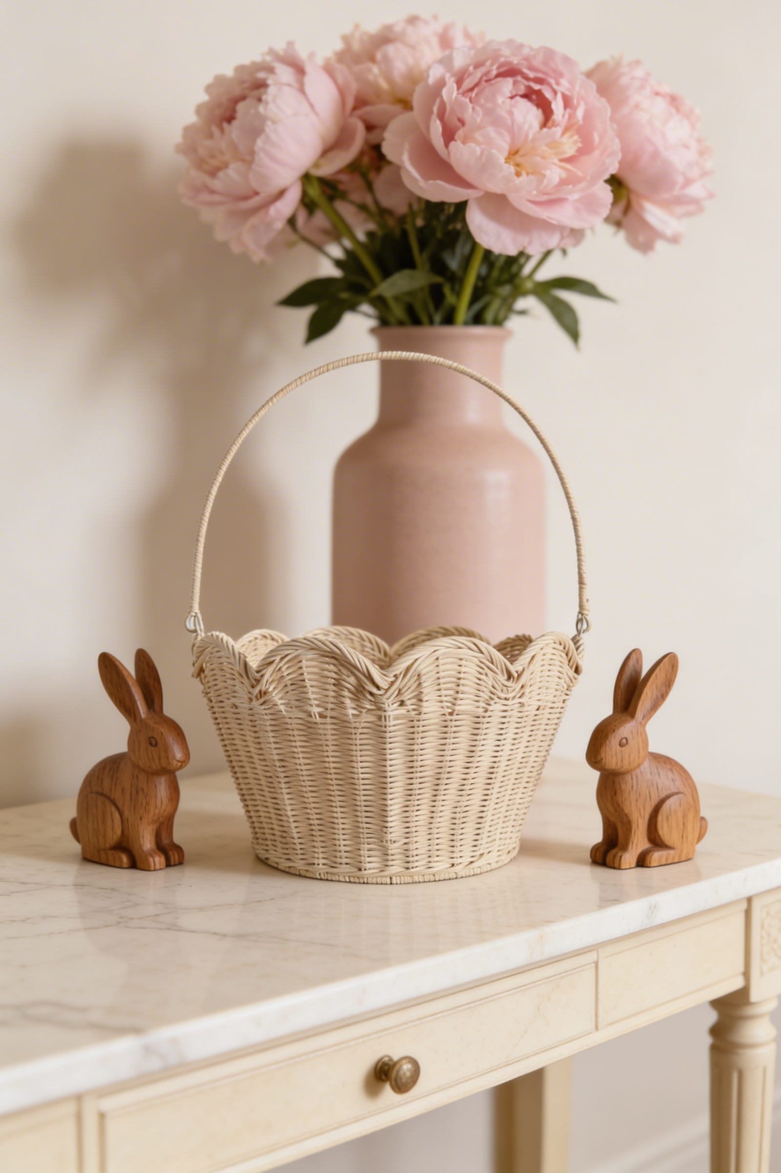 Scallop-edged wicker Easter basket on a marble console with wooden bunny figurines and pink peonies in a blush vase