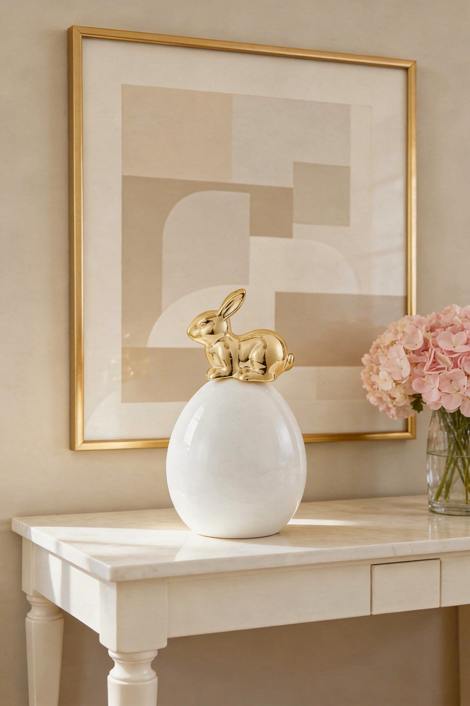 White ceramic egg with gold rabbit figurine on top displayed on a marble console with a gold-framed art print and pink hydrangeas