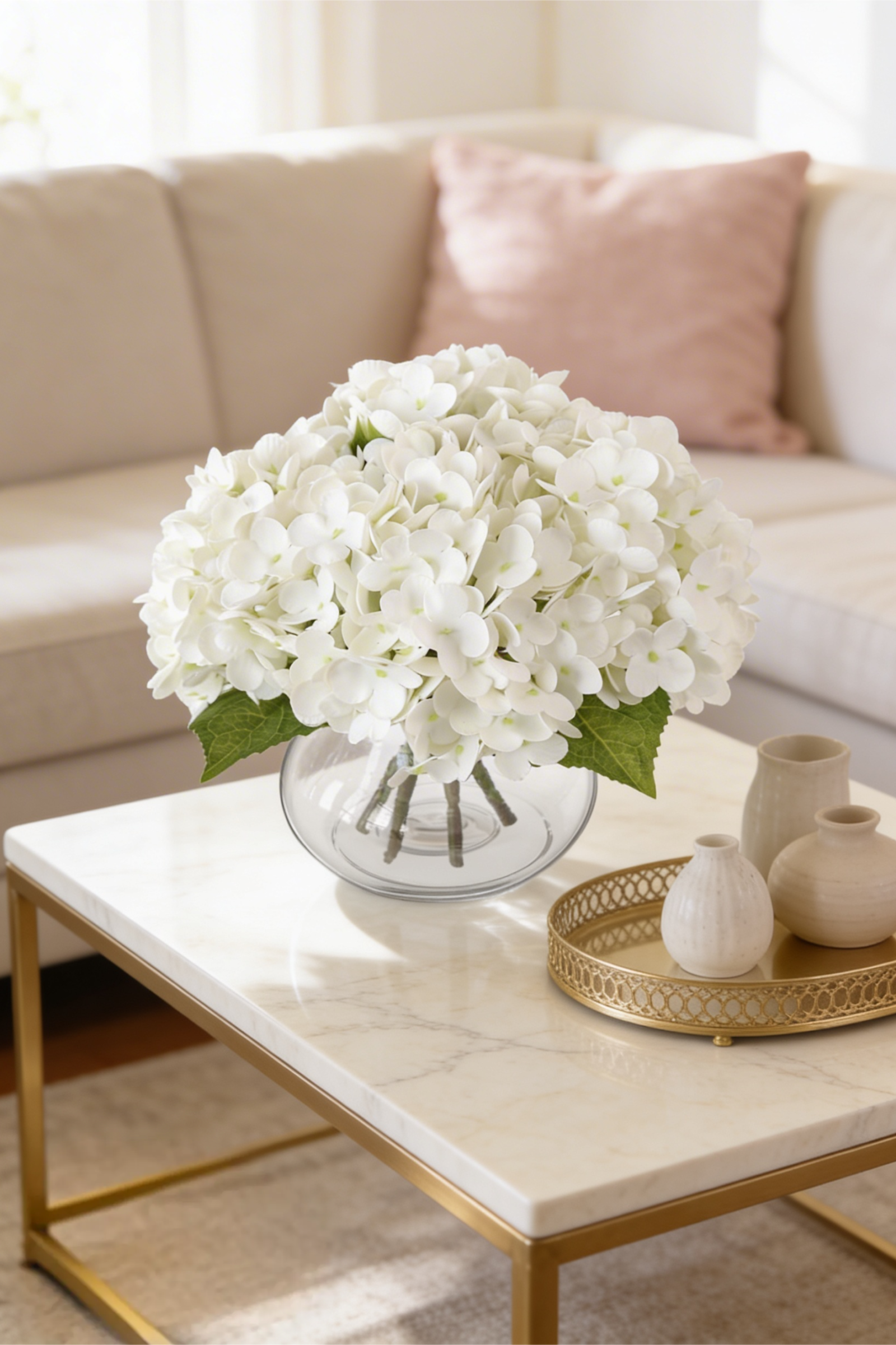 Generous arrangement of white hydrangeas in a glass bowl vase on a marble coffee table with small ceramic vases on a gold tray