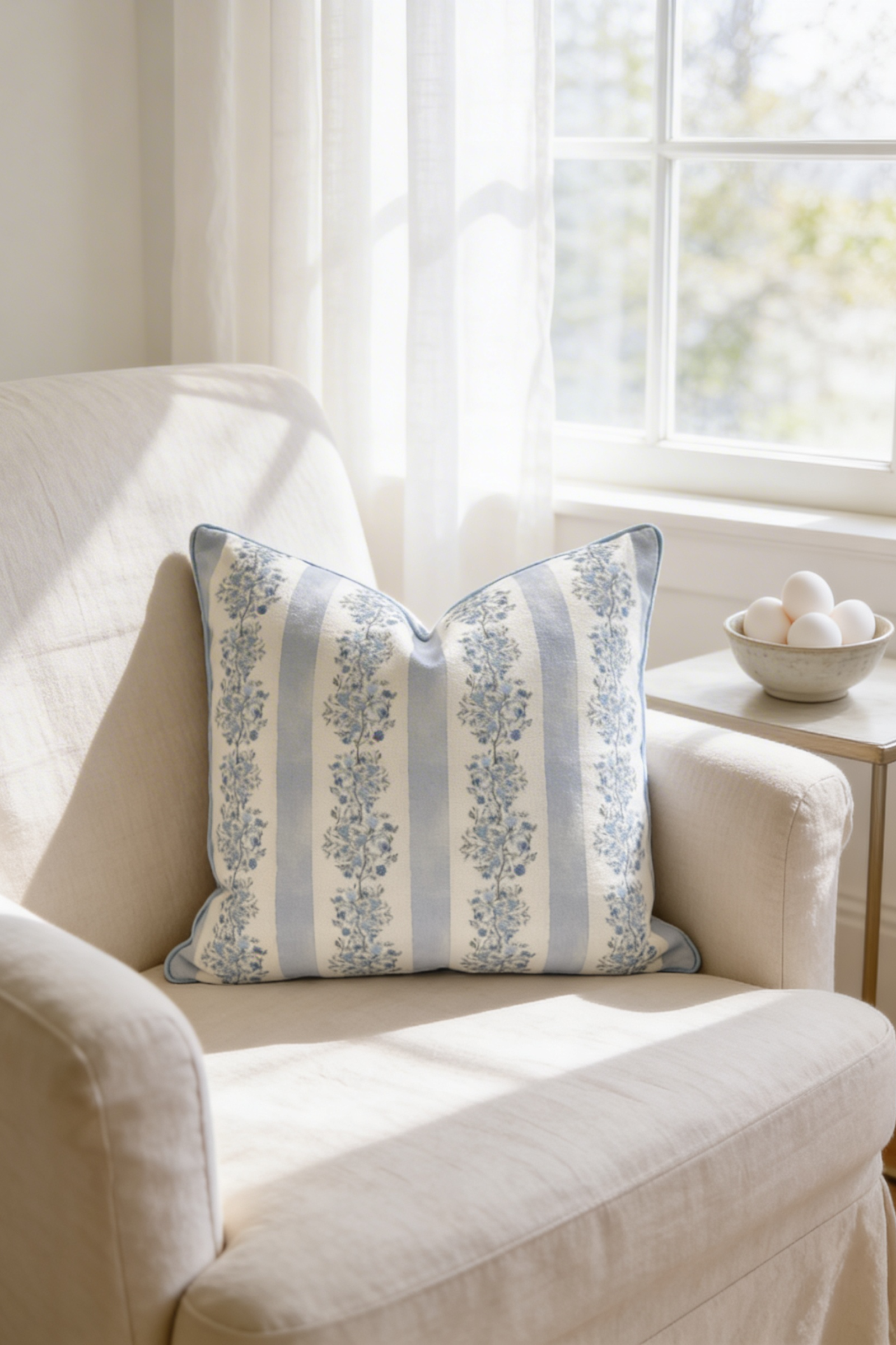 Blue and cream floral stripe throw pillow on a linen armchair by a sunlit window